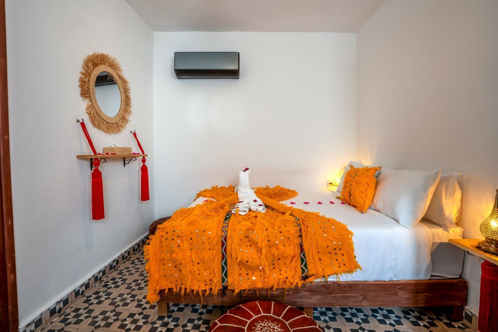 A room with a bed, a wall-mounted air conditioner, a mirror, and a shelf with red tassels.