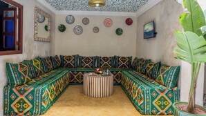 Quarto com sofá estofado em padrão verde e laranja, uma mesa de centro redonda e um vaso com planta.