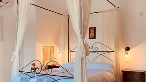 Una cama con dosel, cortinas blancas, una mesita de noche con lámpara y una mesita auxiliar de madera.