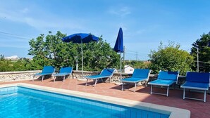 Piscina con bordes de azulejos azules, rodeada de sillones de playa y sombrillas, frente a un cielo despejado.