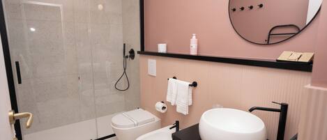 A modern bathroom with a glass-enclosed shower, a white toilet, a bidet, a large round sink, and a mirror on the wall.