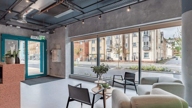 Een moderne lobby met een receptiebalie, een zithoek en grote ramen met uitzicht op de straat en gebouwen.