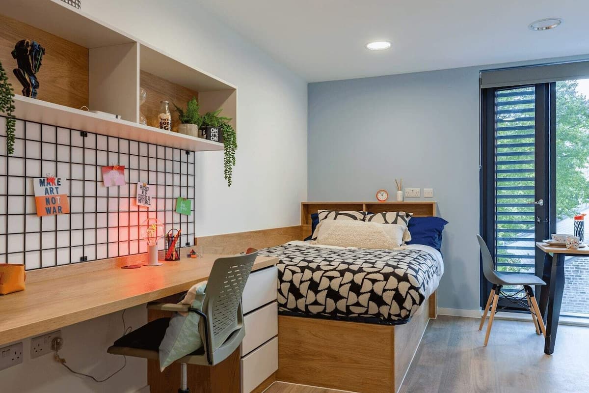 A modern bedroom with a wooden desk, a bed with patterned bedding, and a view of greenery through the window.