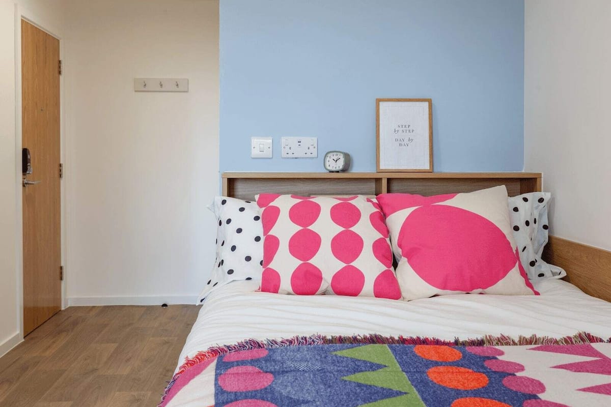 A bedroom with a bed, a wooden door, a light blue wall, a framed quote, a clock, and a power outlet.