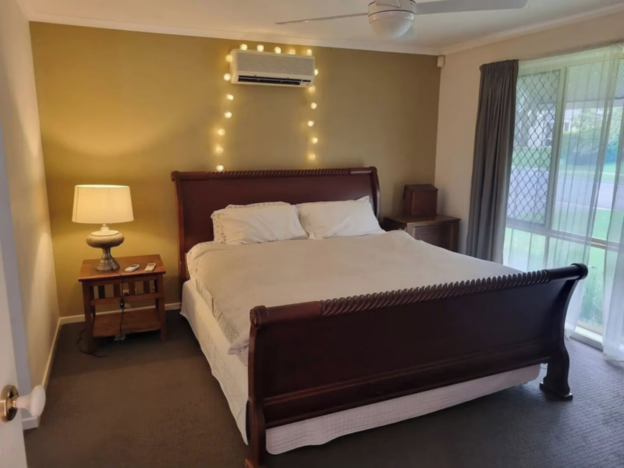 Un dormitorio con una cama grande, una mesita de noche con lámpara, una mesita auxiliar y un ventanal con cortinas.