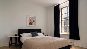 A bedroom with a bed, a bedside table, a small round table, a window with black curtains, and a framed picture on the wall. - Maison Rasco • One-Bedroom Apartment • Old Montreal • Central • Self Check-In Access • Full Kitchen • Pet Friendly • Wi-Fi • AC (Montreal)