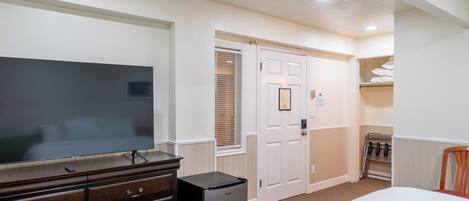 A hotel room with a large flat-screen TV, a dresser, a mini-fridge, a bed, a closet, and a coat rack.