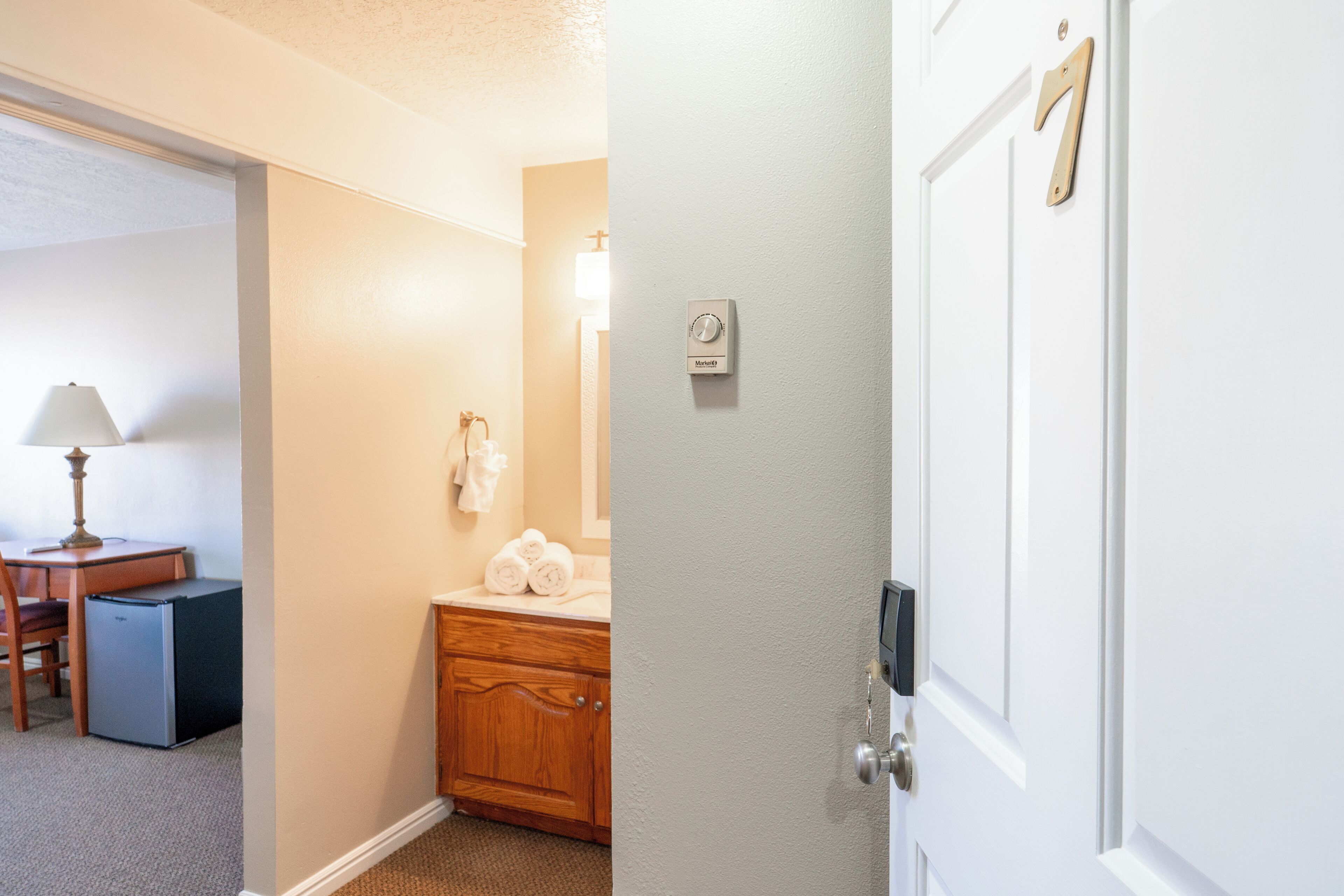 Une chambre d’hôtel avec une commode en bois, un bureau avec une lampe et une salle de bain équipée d’un porte-serviettes.