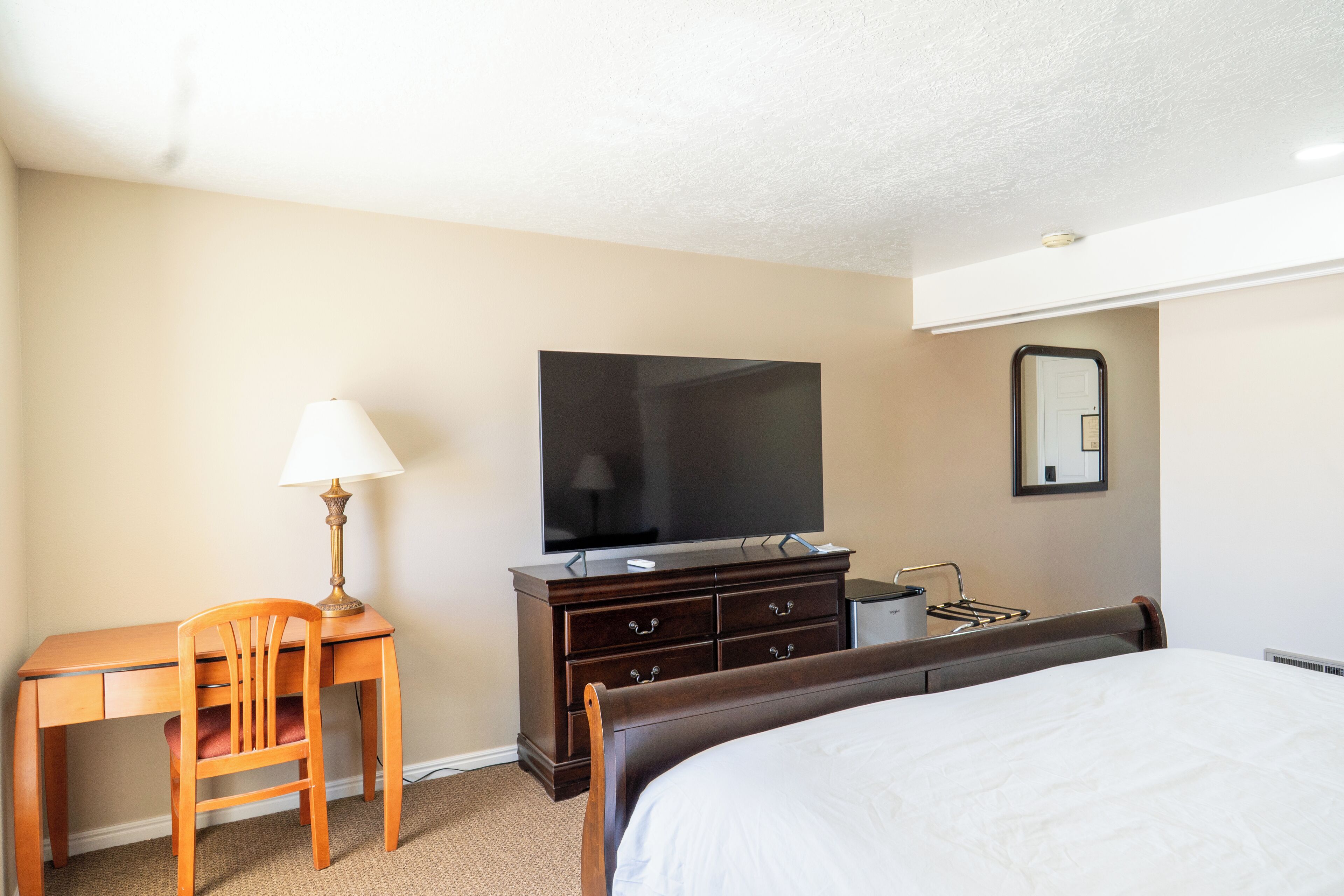 A hotel room with a bed, a desk with a lamp, a dresser, a TV, and a mirror.