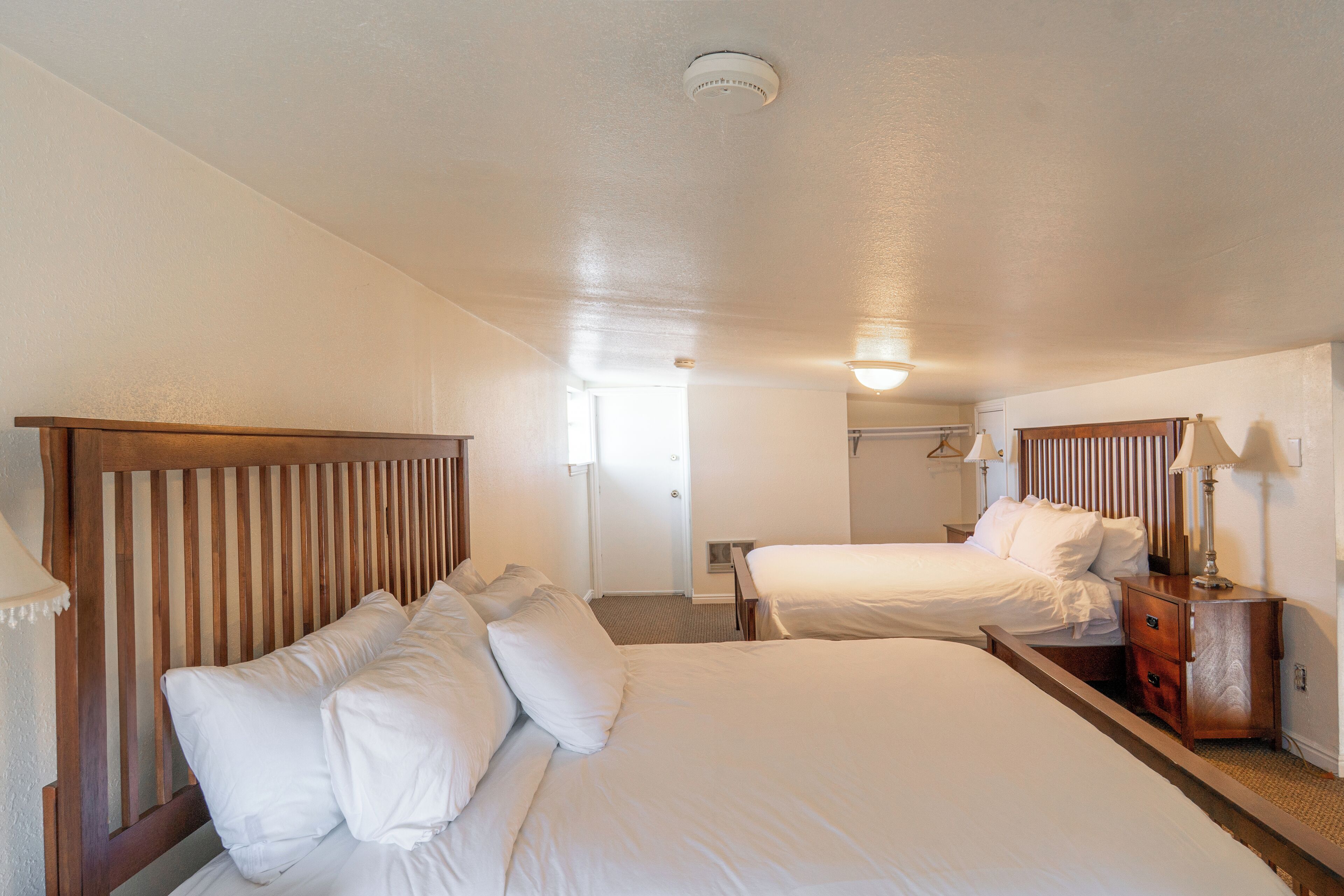 Une chambre d’hôtel avec deux lits, des têtes de lit en bois, du linge de lit blanc, une table de chevet avec une lampe et un luminaire au plafond.