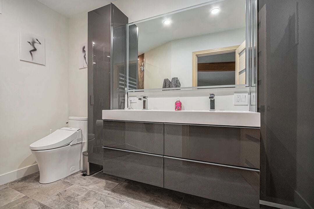 Ein modernes Badezimmer mit Toilette, einem großen Spiegel und einem dunkel gefärbten Waschtisch mit Waschbecken und Arbeitsplatte.