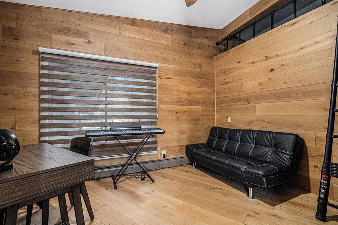 A room with wooden walls, a black leather sofa, a piano, and a table with a fan.