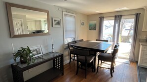 Un comedor con una mesa y sillas de madera, un banco con una planta y una vela, un espejo grande y vistas al exterior a través de una ventana con cortinas.