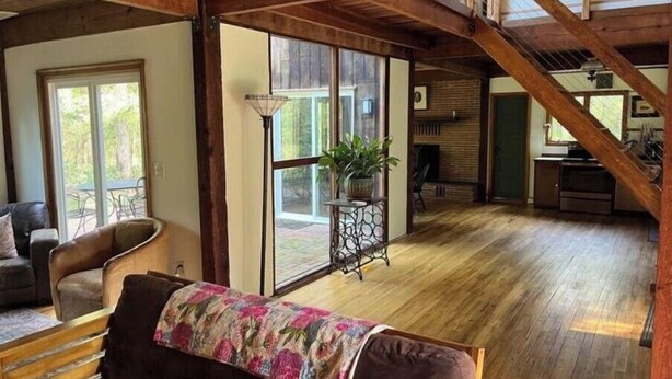 Amplia sala de estar con piso de madera, un sofá con una manta floral, una mesa de comedor y una escalera que lleva a un nivel superior.