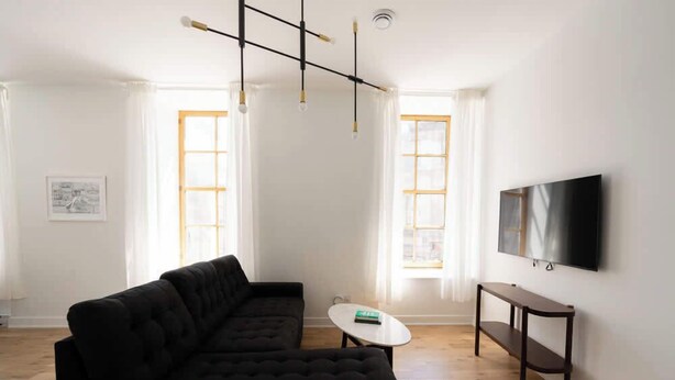 Una sala de estar moderna con un sofá seccional negro, una mesita blanca, un banco de madera y un televisor de pantalla plana montado en la pared.