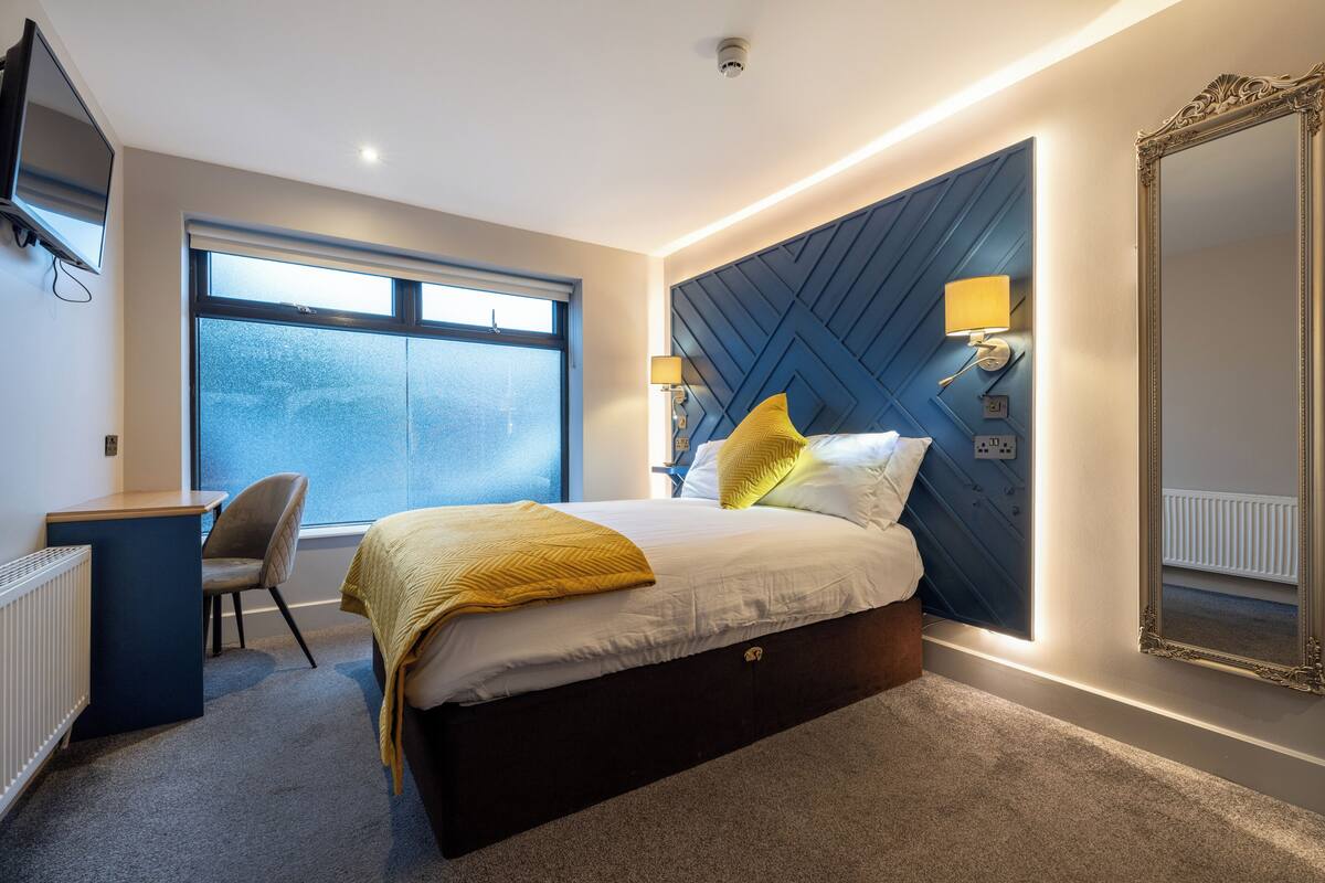 Een moderne hotelkamer met een groot bed, een bureau en een stoel. Een groot raam zorgt voor natuurlijk licht en uitzicht over de stad.