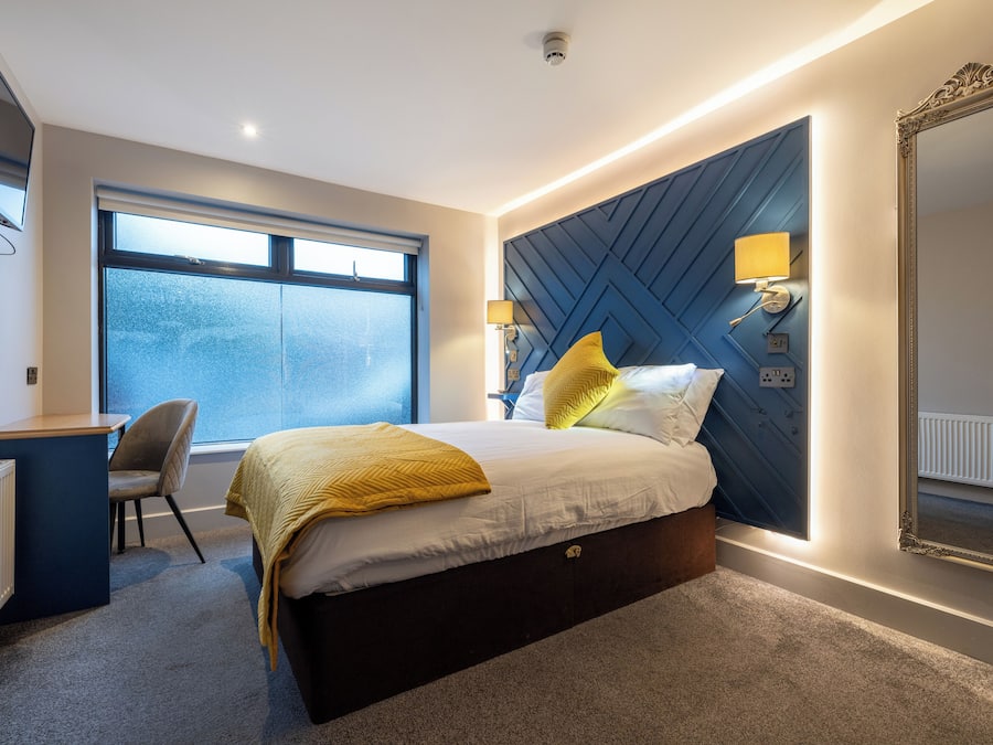 A modern hotel room with a large bed, a desk, and a chair. There is a large window providing natural light and a view of the city.