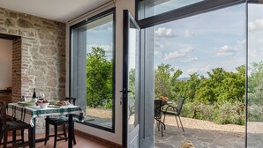 A dining area with a table set for two, a stone wall, and a view of greenery through large glass doors.
