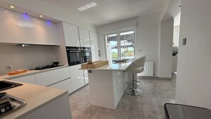 Et moderne køkken med hvide skabe, en central ø med en barstol og udsigt til det fri gennem store vinduer.