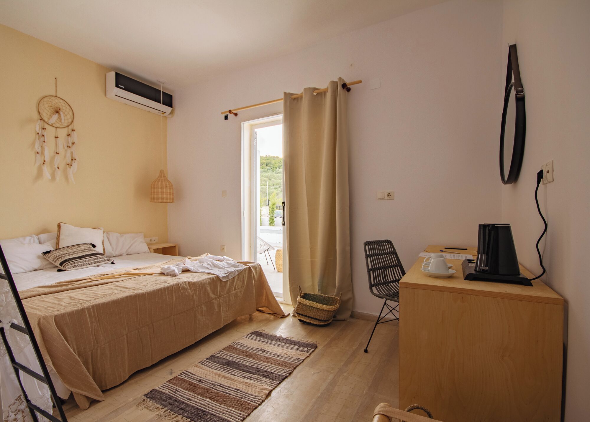 Un dormitorio con cama, mesita de noche y escritorio con silla. Hay una ventana con cortinas, un atrapasueños en la pared y un equipo de aire acondicionado.