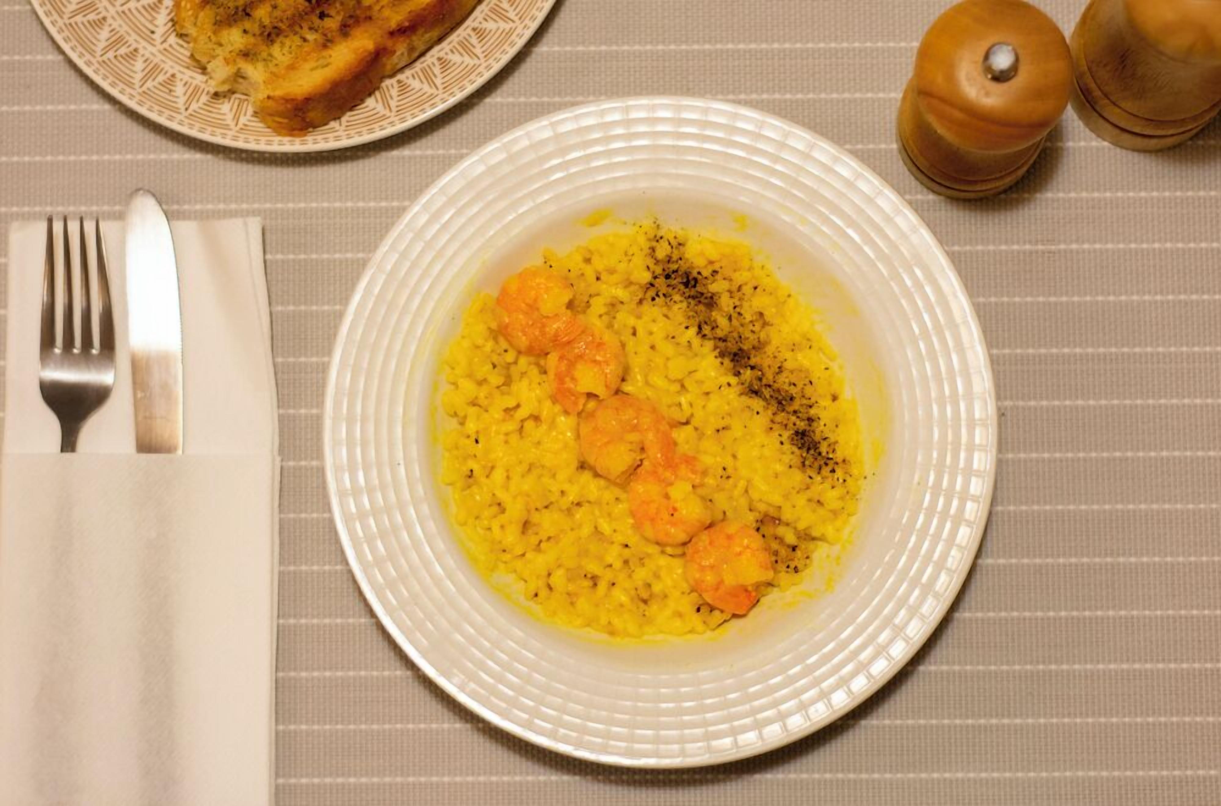 A plate of yellow rice with shrimp and black pepper, a fork, a knife, and a salt shaker on a table.