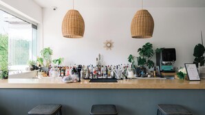 Zona de bar con dos lámparas colgantes de mimbre, una máquina de café y diversos utensilios de bar. Hay tres taburetes y varias plantas en macetas.