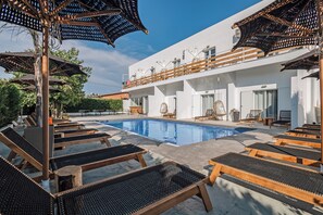 Ein Poolbereich mit Liegestühlen und Sonnenschirmen, ein klares, blaues Schwimmbecken sowie ein weißes Gebäude mit Balkonen.