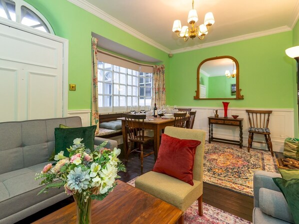 A living room with a sofa, a coffee table with a floral arrangement, a dining table set for four, and a large mirror on the wall.