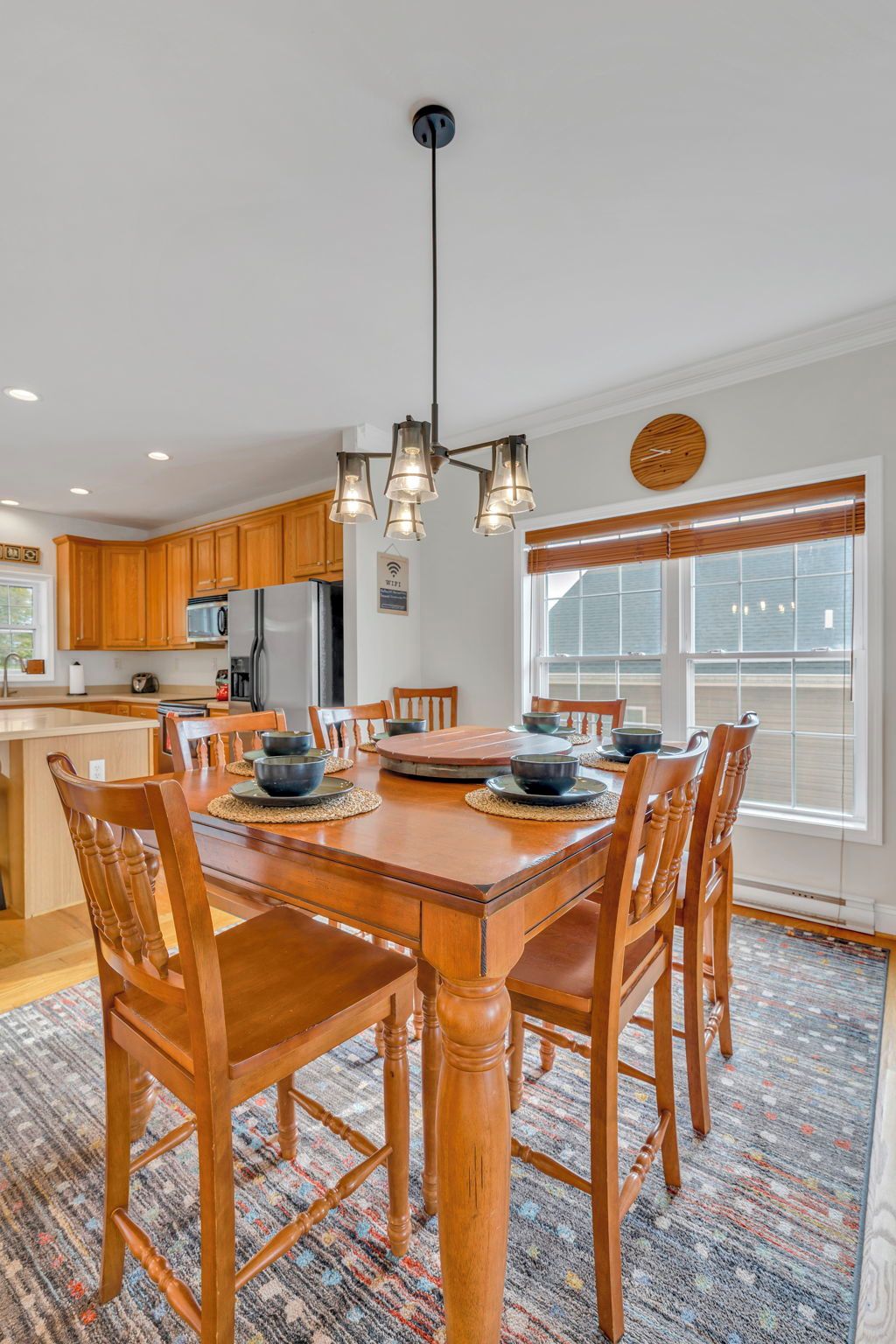 A dining area with a wooden table and chairs, a chandelier, and a kitchen in the background.