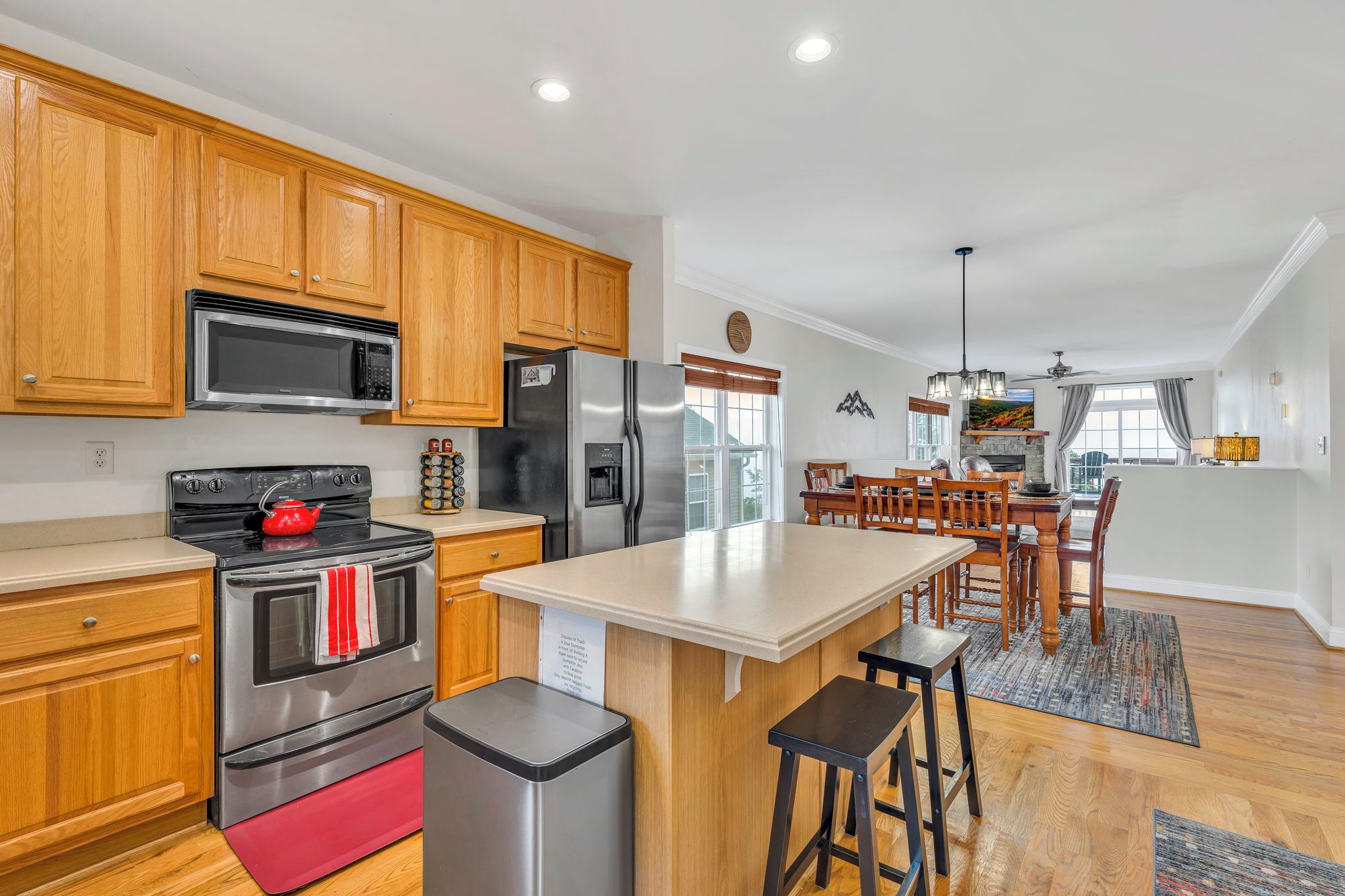 A modern kitchen with wooden cabinets, stainless steel appliances, a central island with stools, and a dining area with a view.