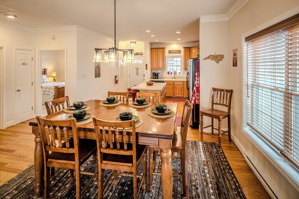 Une salle à manger avec une table et des chaises en bois, une cuisine à l’arrière-plan et une fenêtre avec des stores.