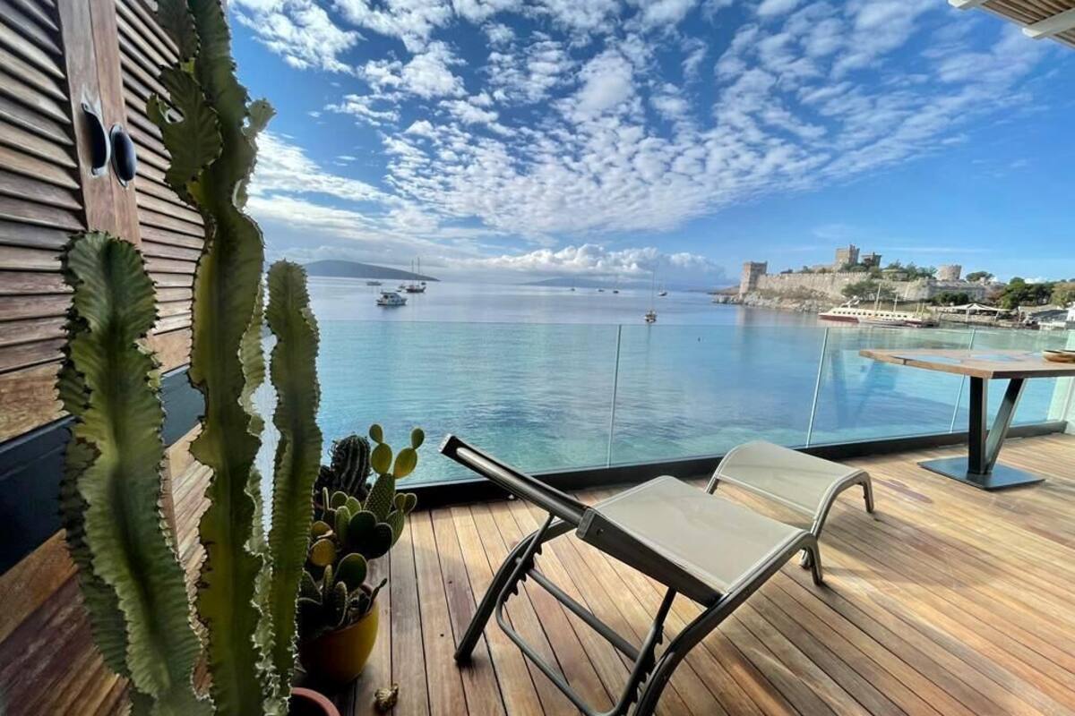 A wooden deck with a metal chair and potted cacti, overlooking a calm sea with boats and a distant castle.
