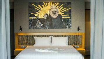 A modern hotel room with a large bed, a headboard with vertical slats, and a wall-mounted artwork depicting a sun and figures.