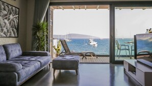 Ein modernes Wohnzimmer mit einer Couch, einem kleinen Tisch und einem Fernseher. Das Zimmer verfügt über eine große Schiebeglasdoor, die auf einen Balkon mit Blick auf das Meer und Boote führt.