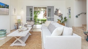 A spacious living room with a white sofa, a coffee table, and a dining table. There is a view of a garden through large windows. - Exclusive one bedroom apartment, a few steps from the Caribbean Sea (Speightstown)
