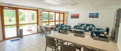 A modern dining area with a long table and chairs, a sofa set, and large windows providing natural light.