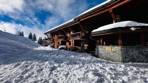 Una cabaña de madera con cimientos de piedra, rodeada de terreno y árboles cubiertos de nieve.