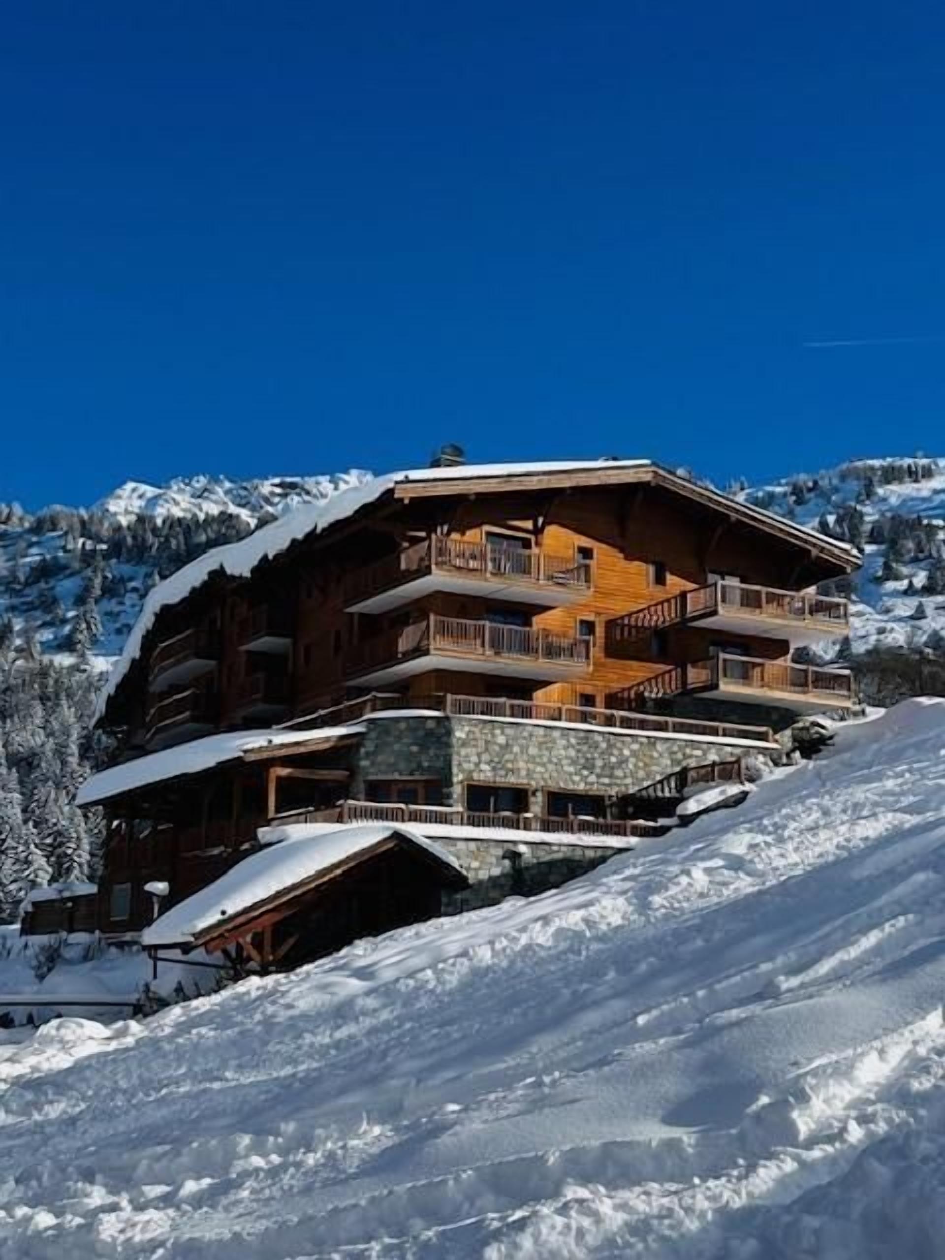 Un chalet en bois à plusieurs étages, doté de balcons, qui se dresse sur fond de montagnes enneigées.