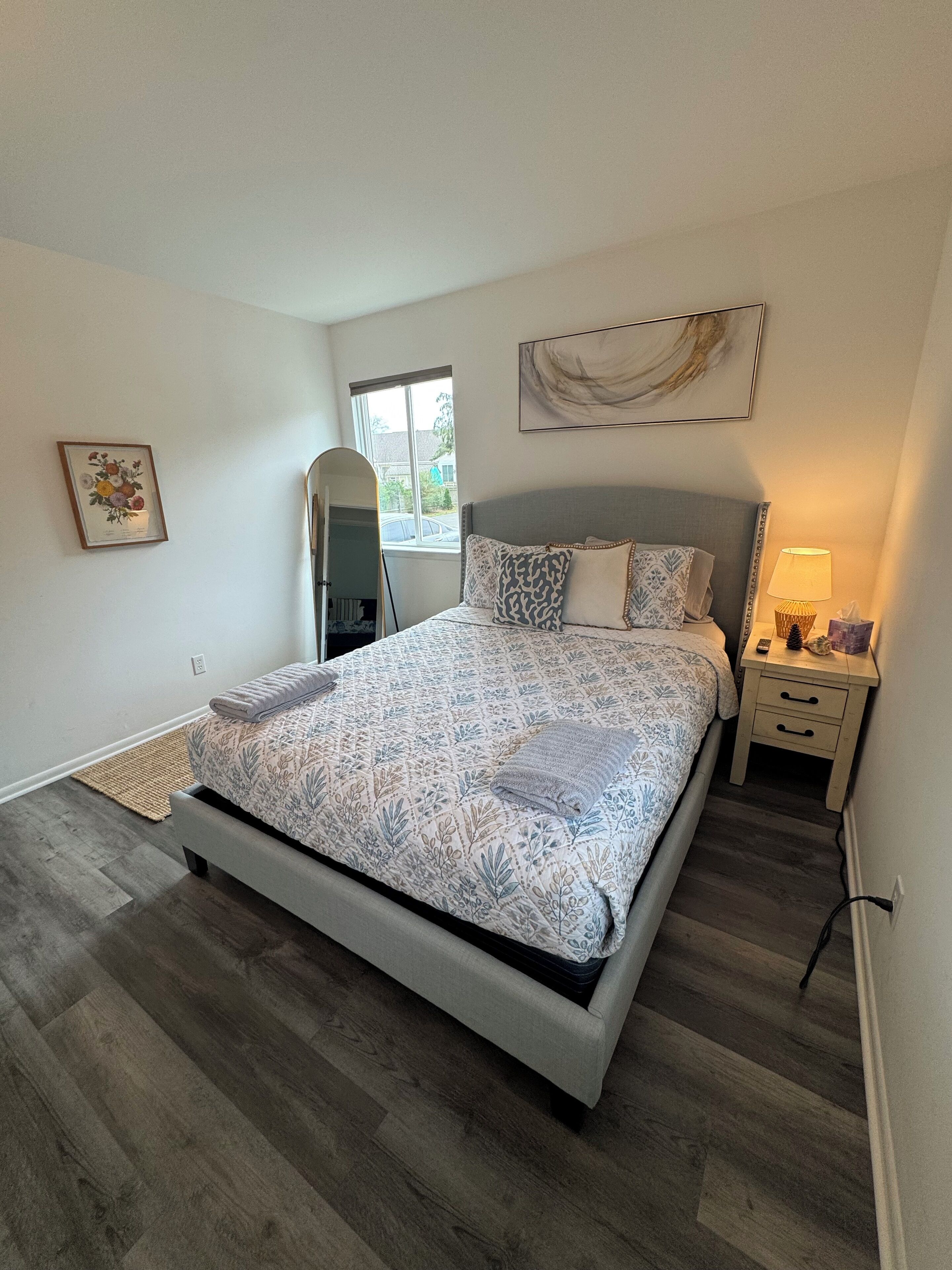 Un dormitorio con una cama grande, una mesita de noche con lámpara, un espejo y una ventana con vistas al exterior.