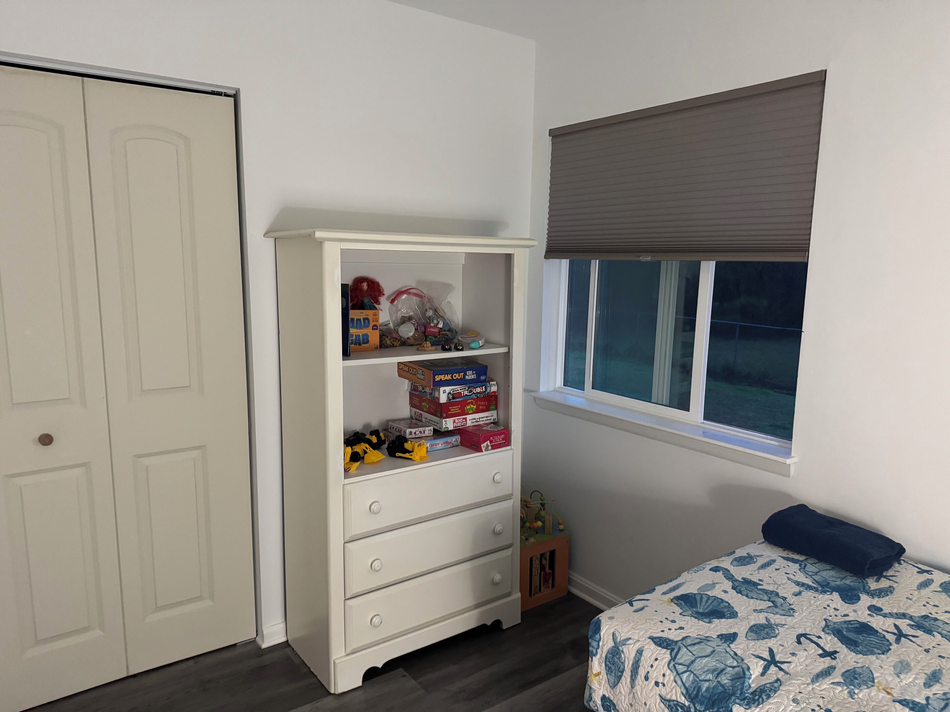 A bedroom with a white dresser, a bed with a blue floral bedspread, and a window with a brown blind.