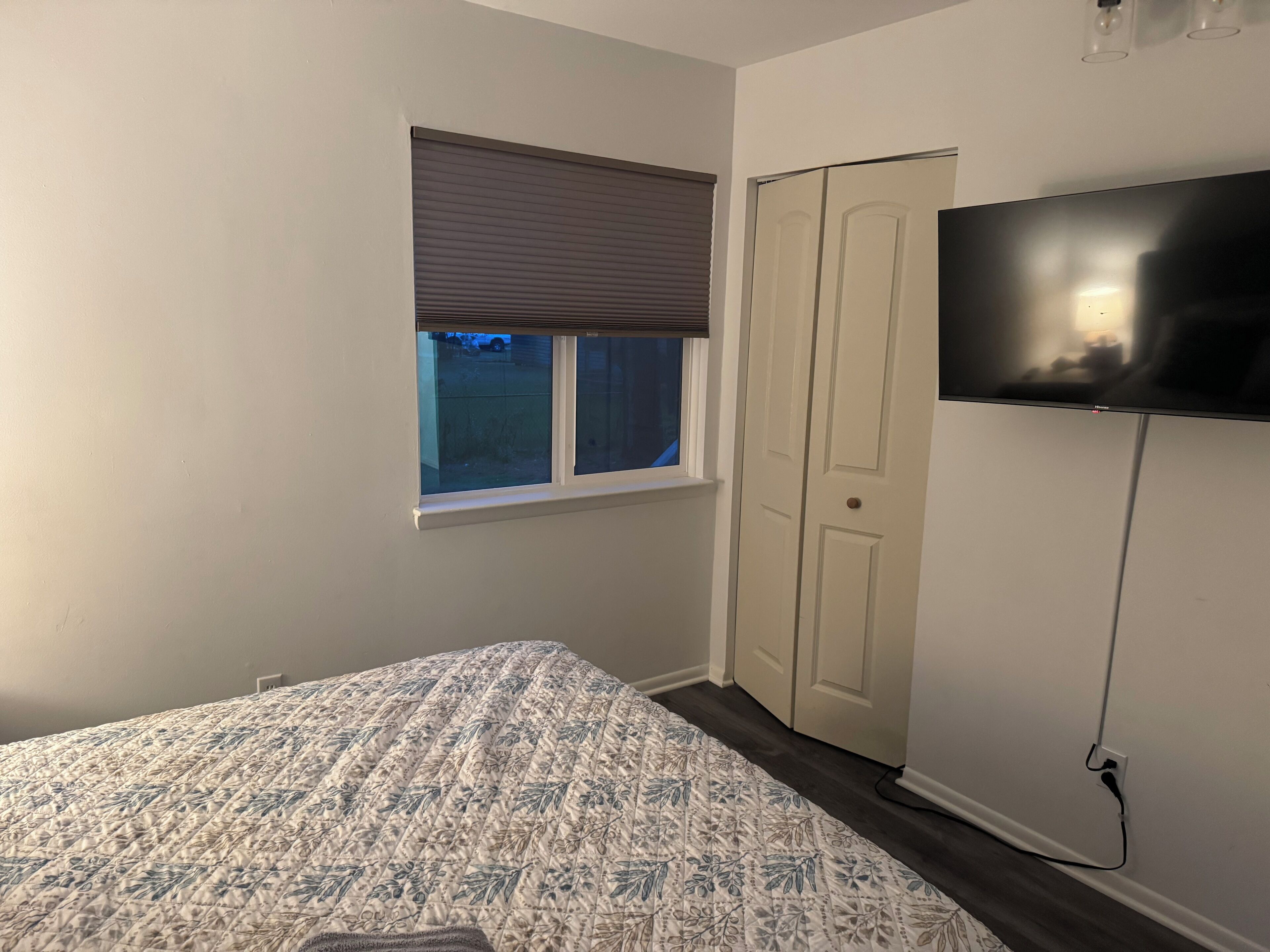 A bedroom with a bed, a window with blinds, a door, and a TV mounted on the wall.
