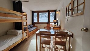 Een knus vertrek met stapelbedden, een houten tafel met stoelen, een bank en een raam met uitzicht op een besneeerd landschap.