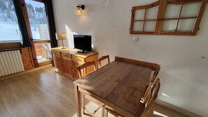 Een eethoek met een houten tafel en stoelen, een televisie op een houten kast, en een raam met gordijnen.