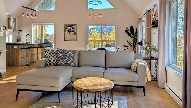 A spacious living area with a large L-shaped sectional sofa, a wooden coffee table, and a dining table with chairs. There are multiple windows providing natural light and views of the outdoors. - Hôtel à la maison - La Villa Foresta (Coaticook)