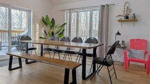 A dining area with a wooden table and benches, large windows, and a red chair. - Hôtel à la maison - La Villa Foresta (Coaticook)