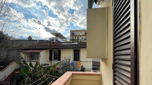 Ein schmaler Balkon mit Fliesenboden, einem geschlossenen Rollladen und Blick auf benachbarte Gebäude und einen teils bewölkten Himmel.