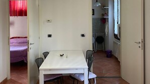 Ein Essbereich mit einem weißen Tisch und Stühlen, einer Hängeleuchte und Blick in eine Küche.