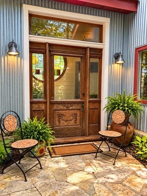 A wooden front door with a glass panel, two wall-mounted lights, and a stone-paved entrance area with two chairs and a potted plant.