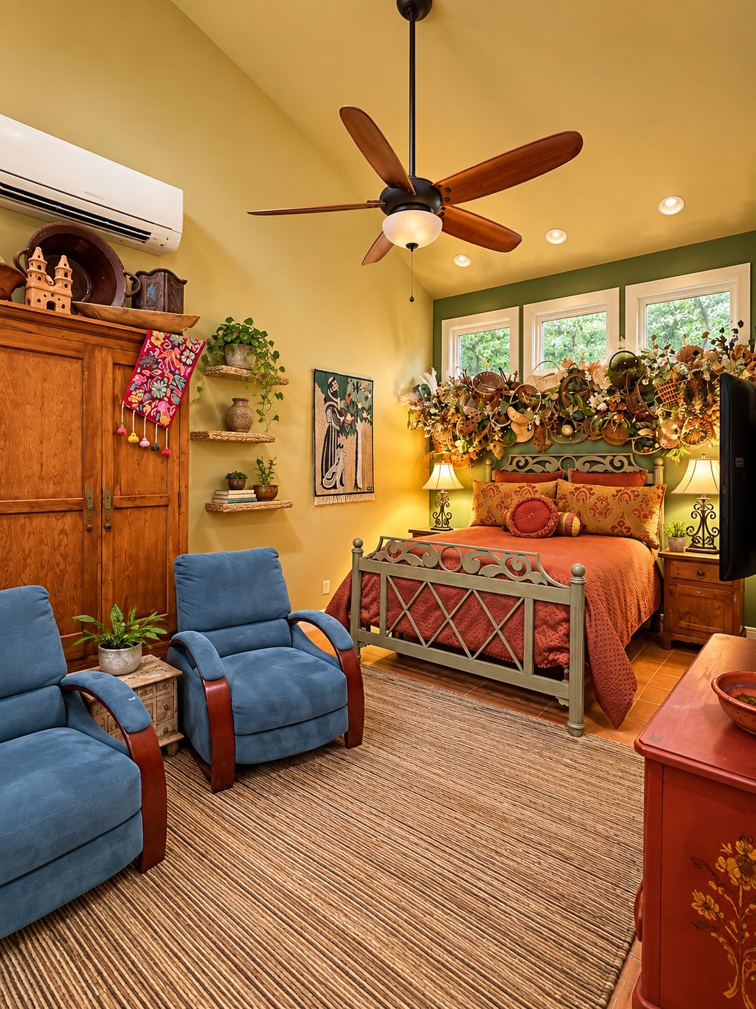 A bedroom with a bed, two blue armchairs, a wooden wardrobe, a ceiling fan, and a window with a view of greenery.