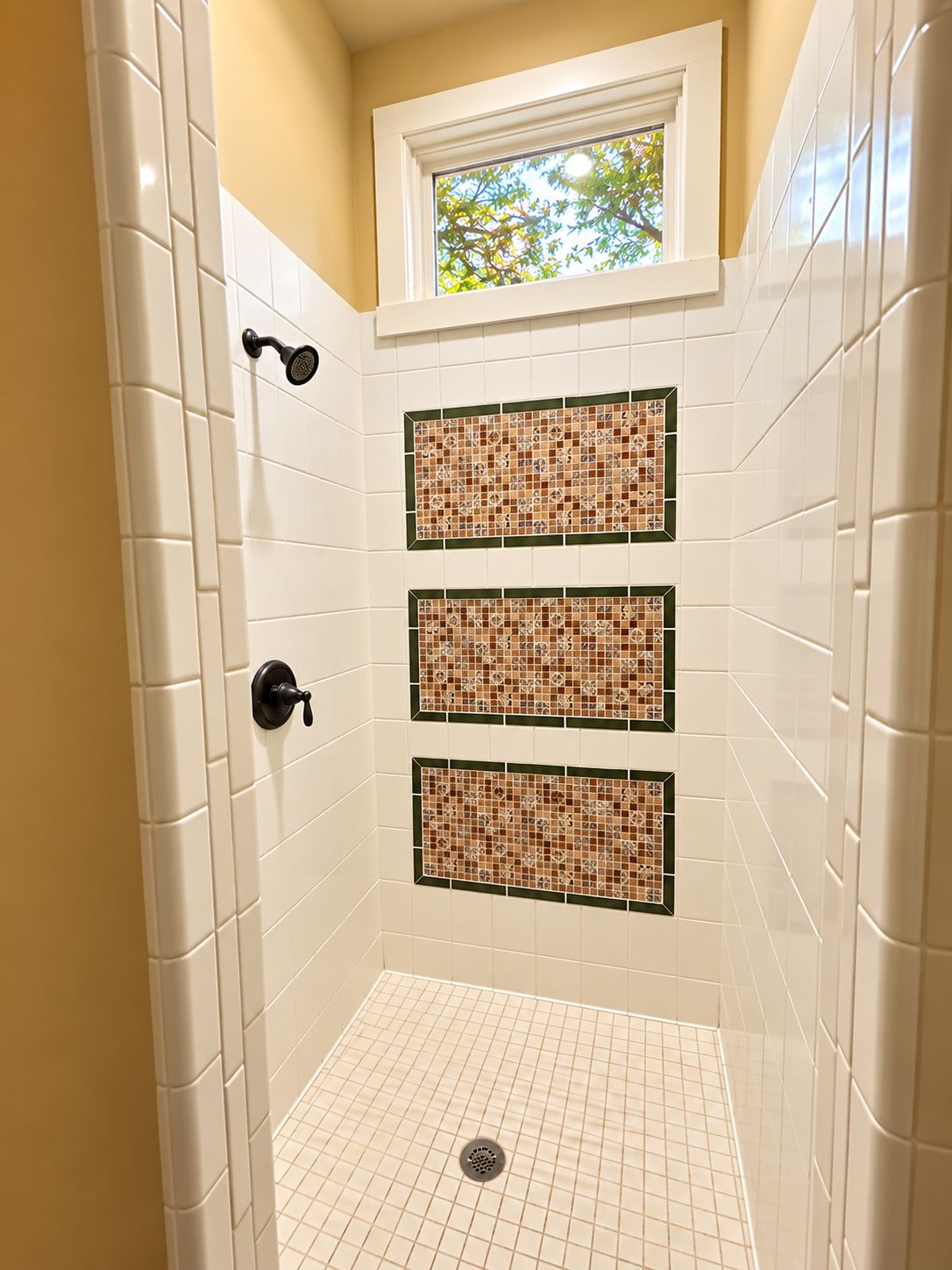 A shower with white tiles, a window, and a decorative mosaic panel.